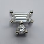 Close-up of two clear faceted stones—an emerald-cut rectangle above and a round brilliant below—set in polished metal prongs.