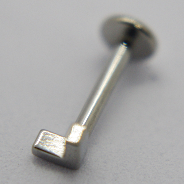 Macro close-up of a small shiny L-shaped silver metal pin with a flat disk back resting on a pale surface.