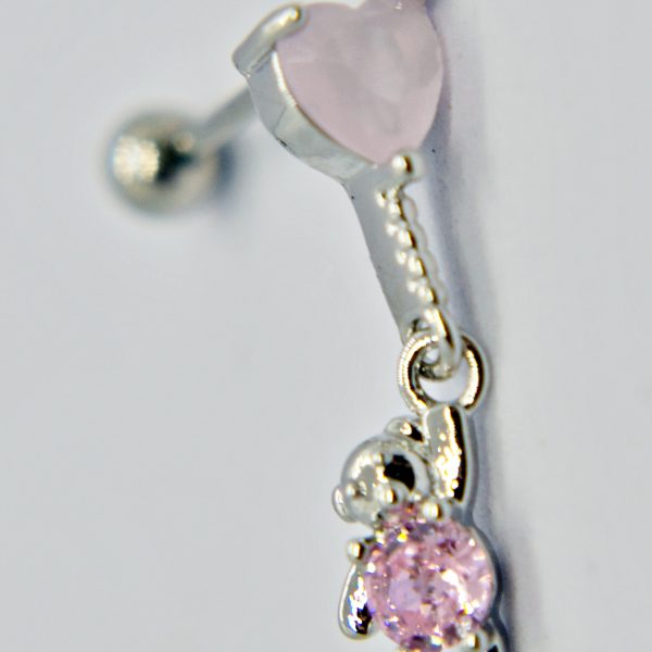 Close-up of a silver earring with a pale pink heart stud and a dangling teddy-bear charm holding a round pink gemstone.