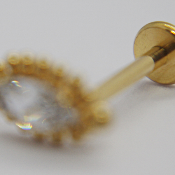 Gold stud earring with marquise-shaped clear gemstone surrounded by tiny gold beads, shallow focus with gold backing visible.