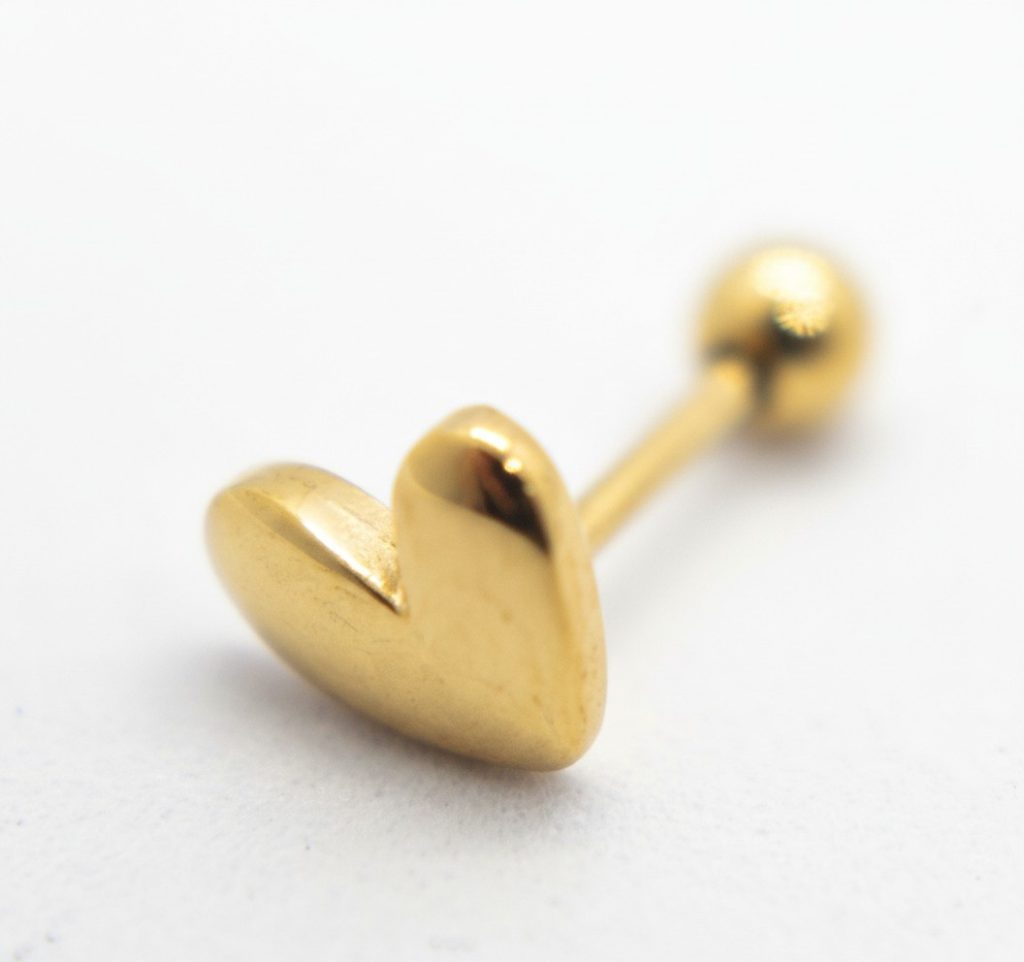 Gold heart-shaped barbell piercing stud with round screw-back resting on a white surface.