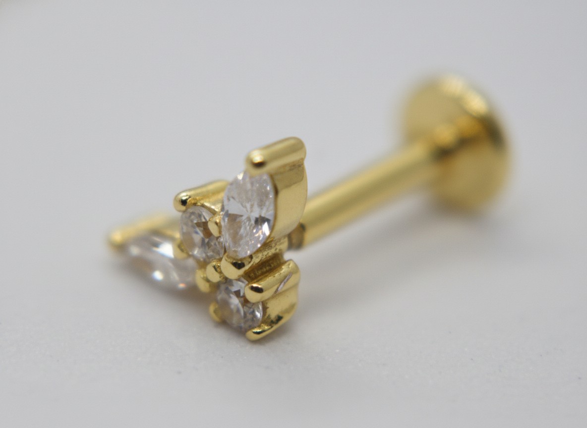 Close-up of a gold labret stud with three clear faceted stones set in a small triangular cluster on a gray surface.