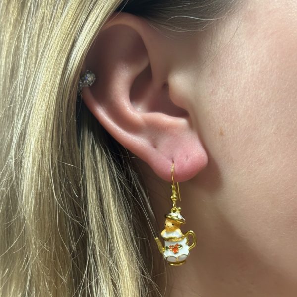 Blonde ear wearing a gold-and-white teapot earring with orange flower detail and a small crystal cartilage stud.