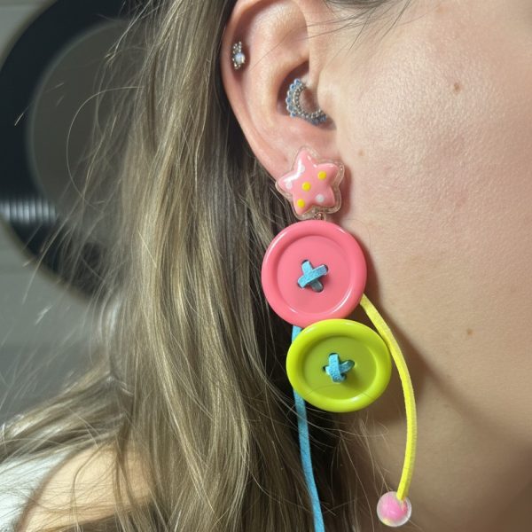 Right ear with multiple piercings wearing a pink star stud and dangling oversized pink and yellow button charms with blue and yellow cords