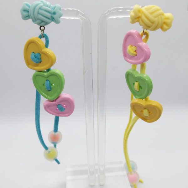 Pair of pastel dangle earrings on a clear stand with candy-shaped studs and three heart-button charms on colored cords.