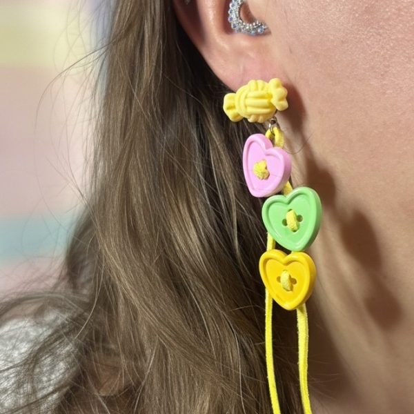 Ear with a rhinestone ear cuff and a long dangle earring featuring a yellow candy-like stud and three pastel heart buttons on yellow cords with beads.