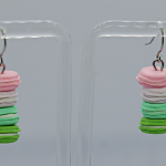 Pair of dangling earrings shaped like stacked pastel macarons — pink, white, mint and green — hanging on a display stand.