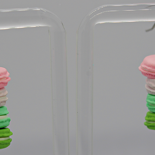 Dangling earrings shaped like stacked pastel macarons (pink, white, green) hanging from silver hooks on a clear stand.