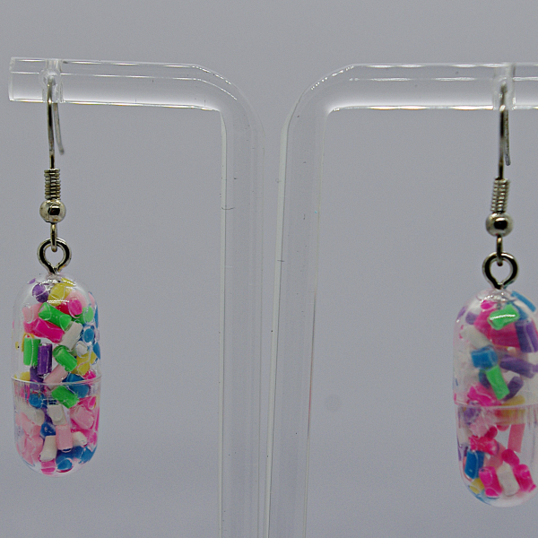 Pair of clear capsule earrings filled with tiny multicolored beads, suspended from silver hooks on a transparent acrylic stand.