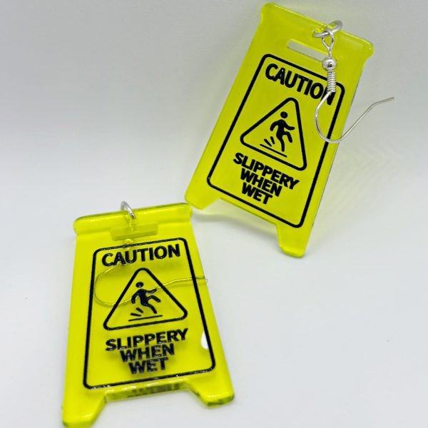 Small neon-yellow caution signs reading Slippery When Wet turned into earrings with silver hooks on a white background.