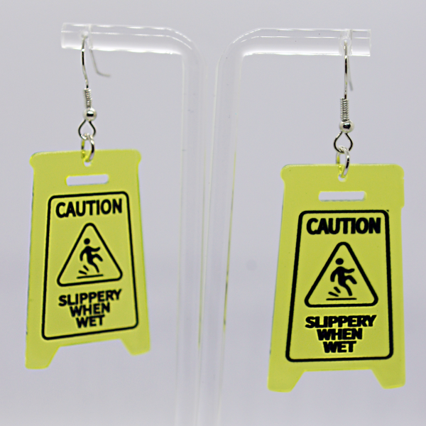 Pair of yellow earrings shaped like miniature wet-floor Caution Slippery When Wet signs hanging from a clear acrylic display.