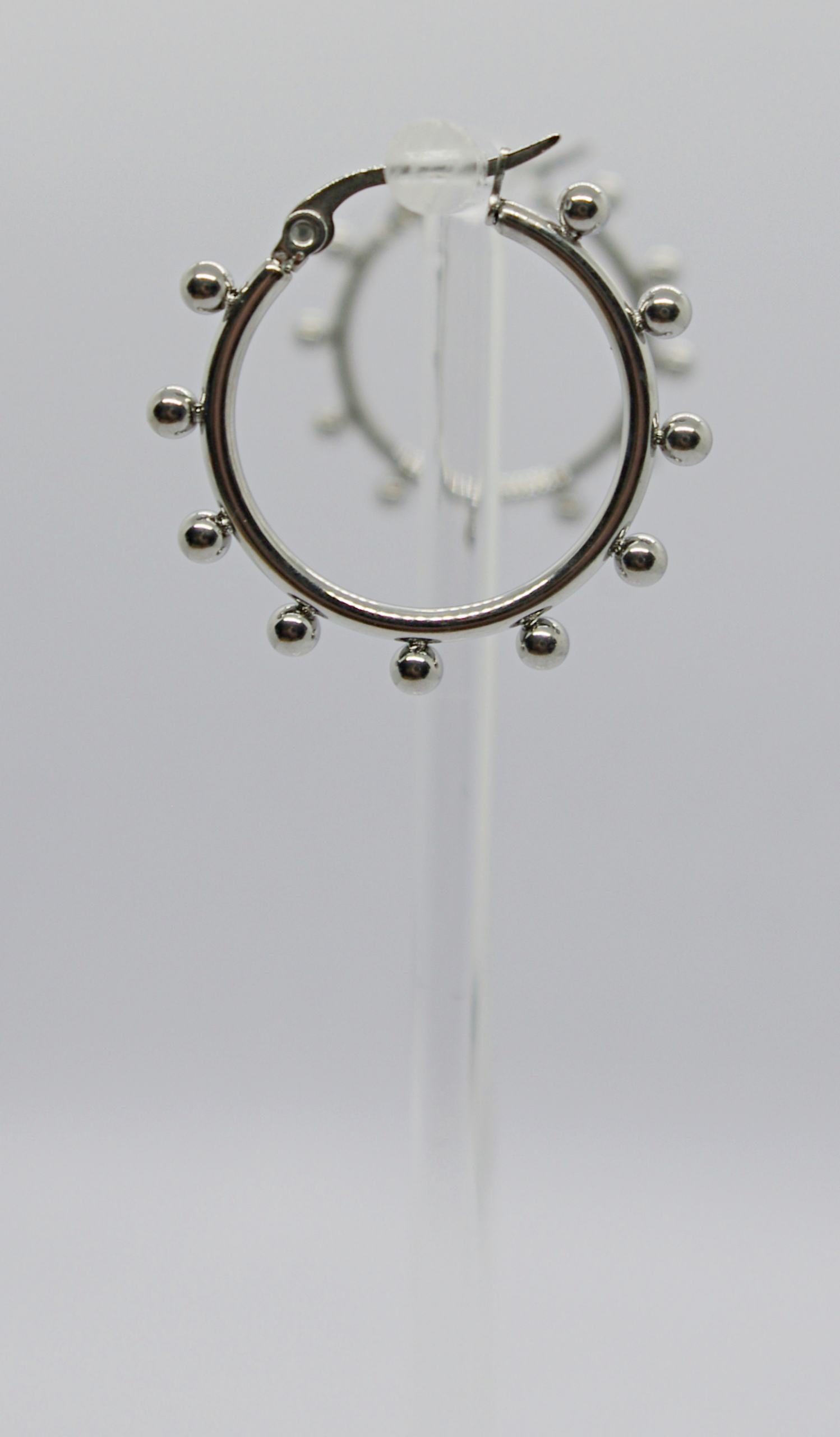 Silver hoop earring with small round beads evenly spaced around it, displayed on a clear stand against a pale background.