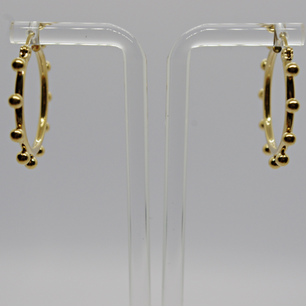 Gold hoop earrings with small round bead accents hanging from a clear acrylic display stand against a soft gray background.