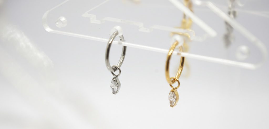 Pair of small hoop earrings on a clear acrylic display, one silver and one gold, each with a marquise-cut clear stone drop.