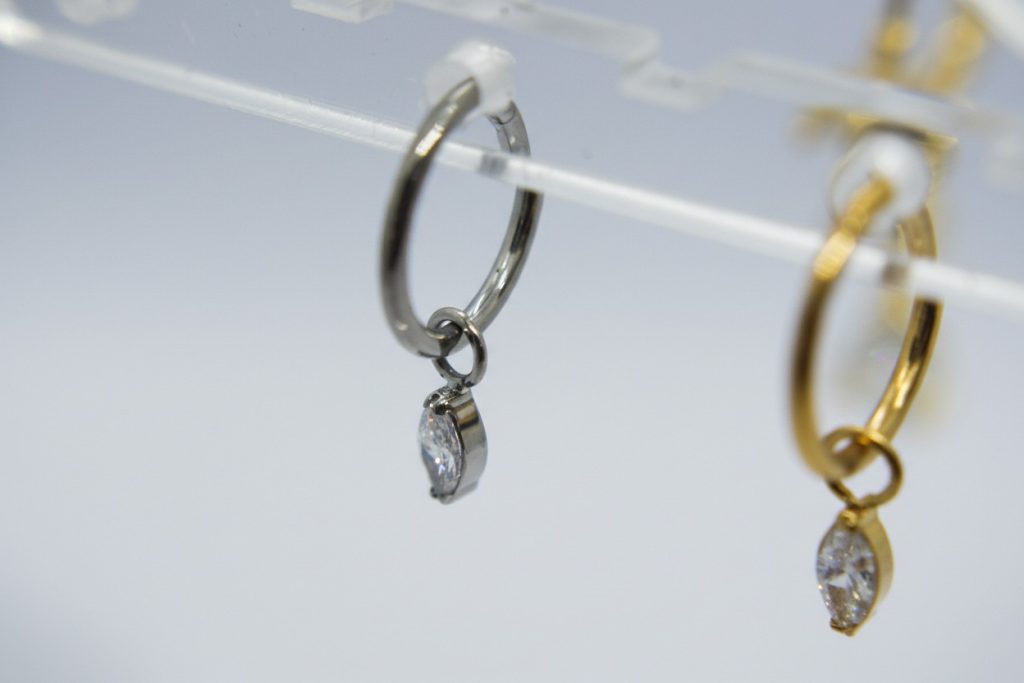 Silver hoop earring with a small marquise-cut clear stone charm hanging on a display, a blurred gold hoop beside it.