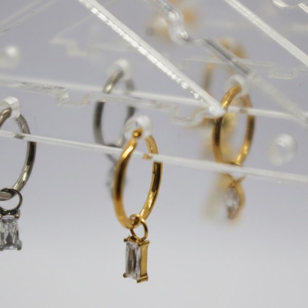 Silver and gold small hoop earrings hanging on a clear acrylic rack, each with a rectangular baguette-cut drop stone.