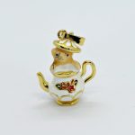 Small white enamel teapot charm with gold trim and red floral detail, topped by a tiny figurine, on a plain white background.