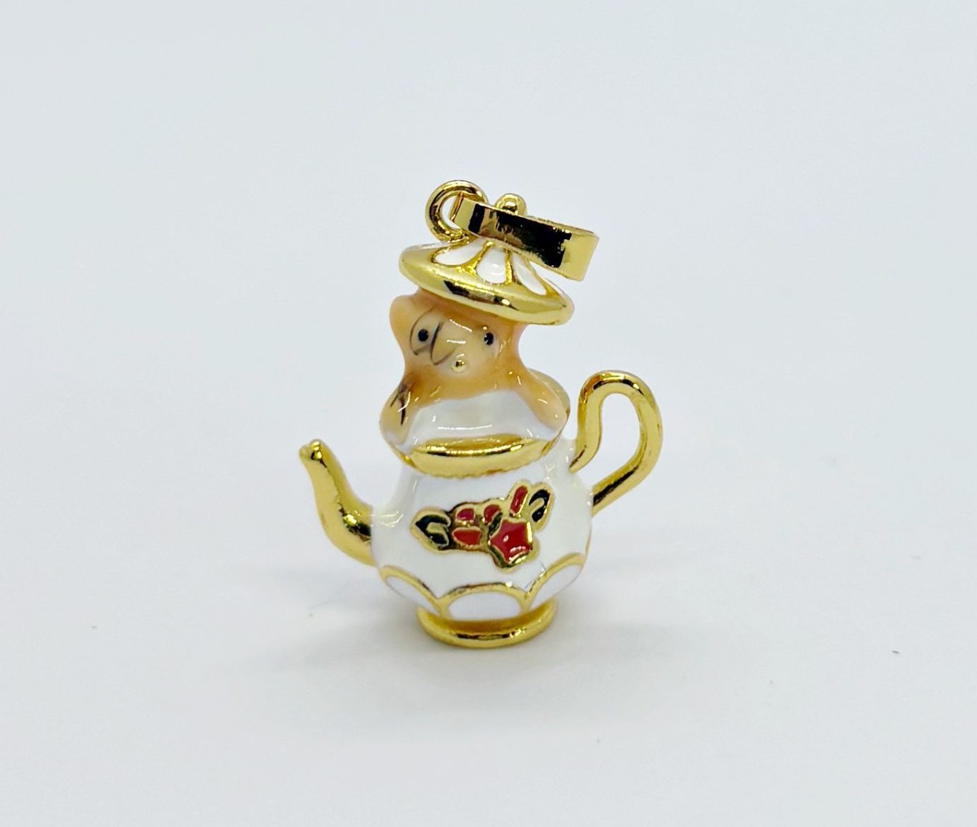 Small white enamel teapot charm with gold trim and red floral detail, topped by a tiny figurine, on a plain white background.