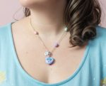 Silver chain with pastel beads and a small lavender heart locket pendant with glittery multicolored inlay at a woman's collarbone.
