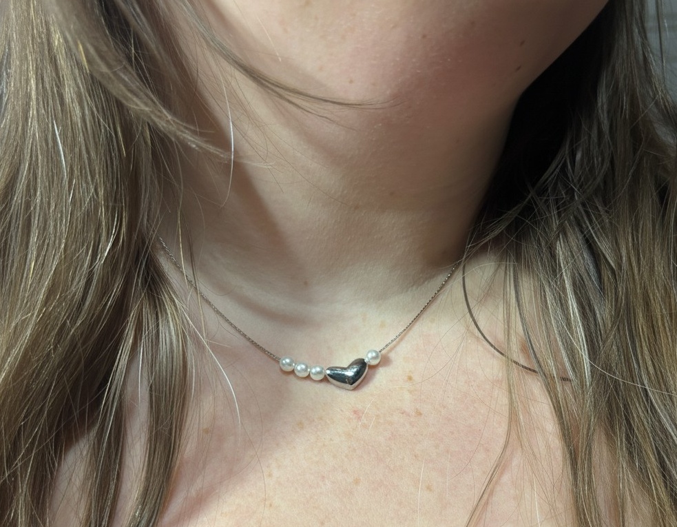 Close-up of a neck wearing a delicate chain with five small pearls and a silver heart pendant, hair at the sides.