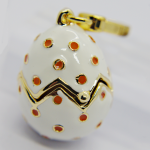 White enamel egg-shaped charm with gold zigzag band and orange polka dots, topped by a gold clasp