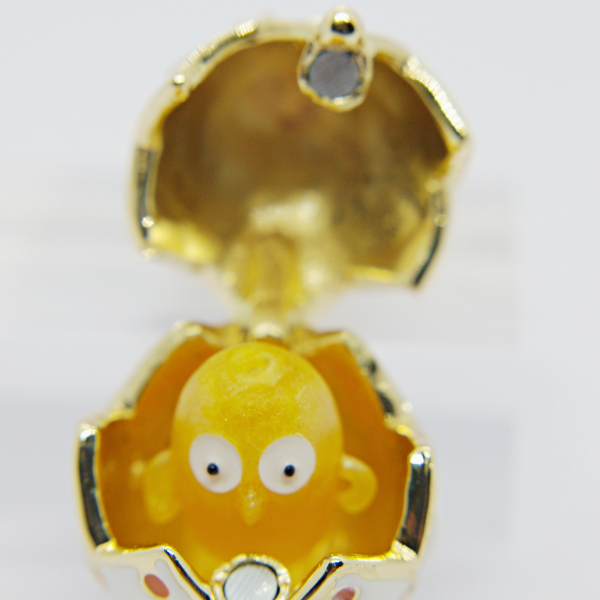 Small yellow toy chick with wide white eyes peeking from a white-and-gold decorative cracked egg with hinged lid.