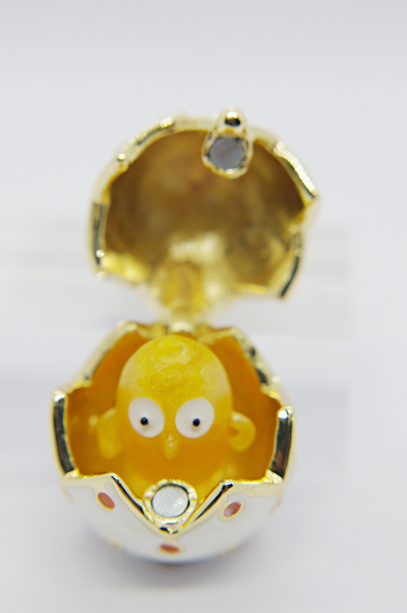 Small yellow toy chick with wide white eyes peeking from a white-and-gold decorative cracked egg with hinged lid.