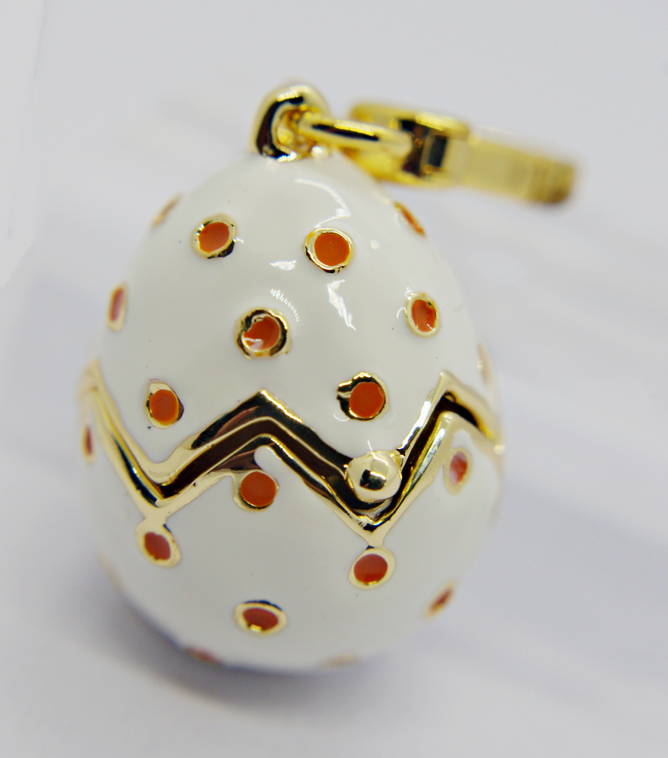 White enamel egg-shaped charm with gold zigzag band and orange polka dots, topped by a gold clasp