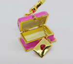 Small pink enamel trinket box with gold trim, open to reveal a dangling gold envelope charm stamped with a red heart.