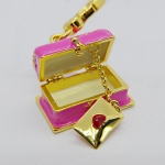 Small pink enamel trinket box with gold trim, open to reveal a dangling gold envelope charm stamped with a red heart.