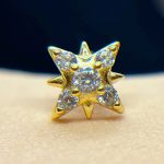 Small gold star-shaped stud earring set with six tiny round clear gemstones, photographed close-up on skin with dark background