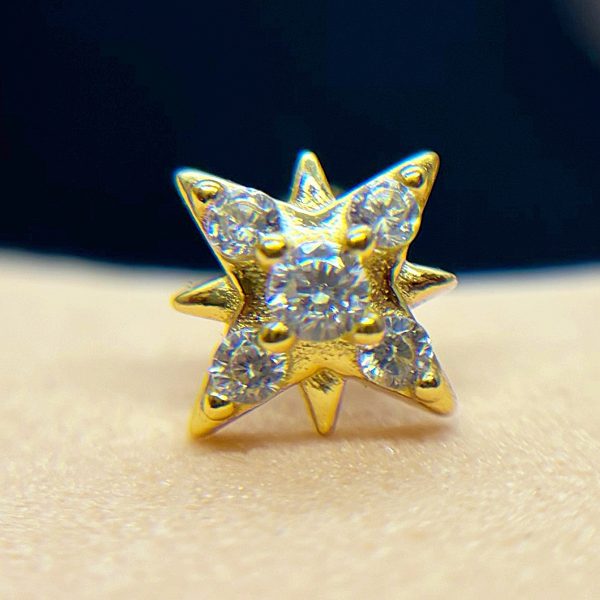 Small gold star-shaped stud earring set with six tiny round clear gemstones, photographed close-up on skin with dark background