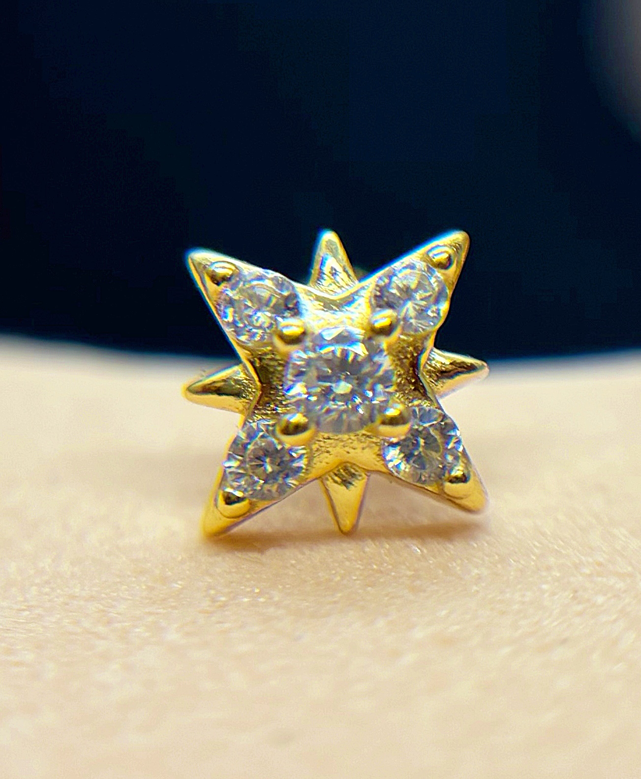 Small gold star-shaped stud earring set with six tiny round clear gemstones, photographed close-up on skin with dark background