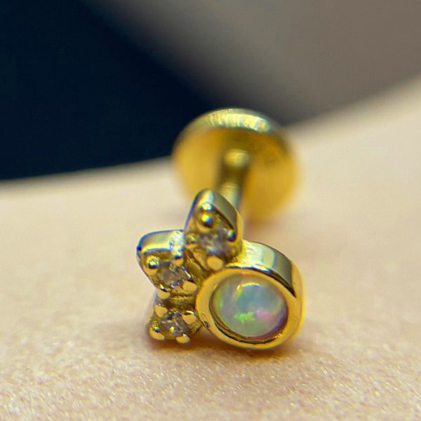 Small gold stud earring with a round iridescent opal and three tiny clear stones arranged like a cluster on skin background.