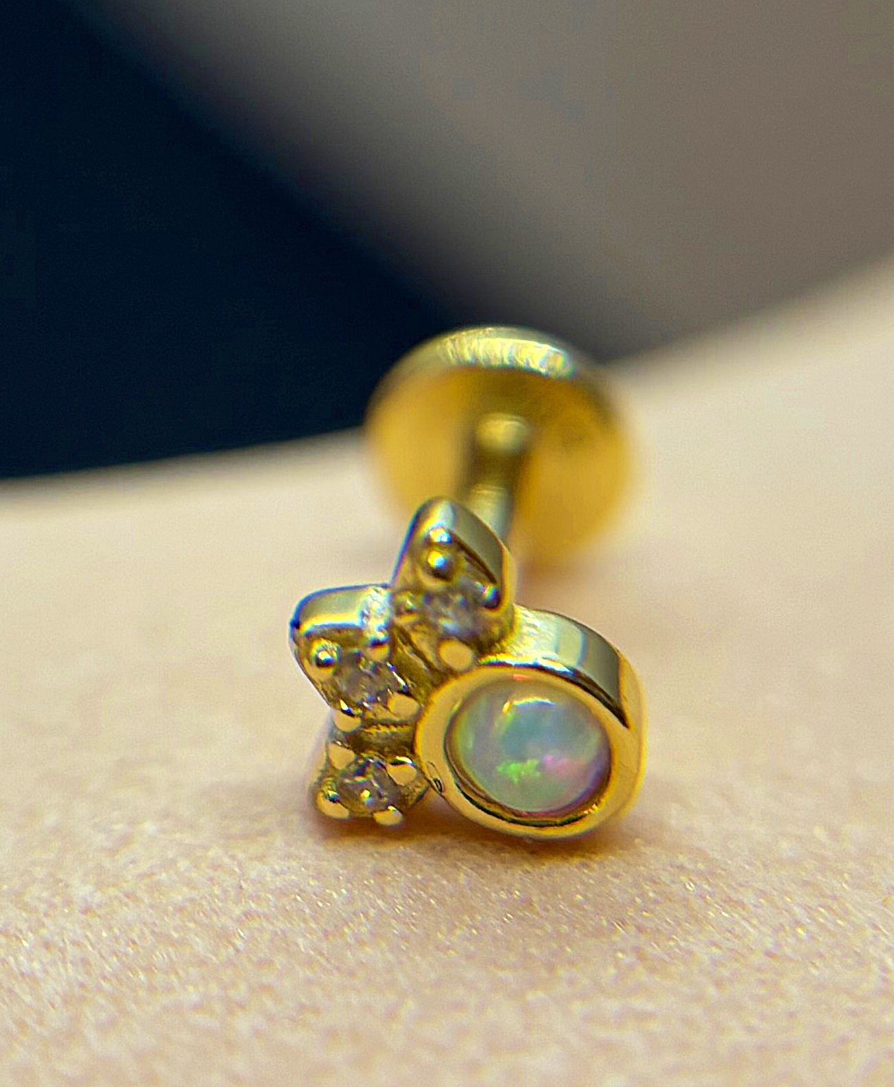 Small gold stud earring with a round iridescent opal and three tiny clear stones arranged like a cluster on skin background.