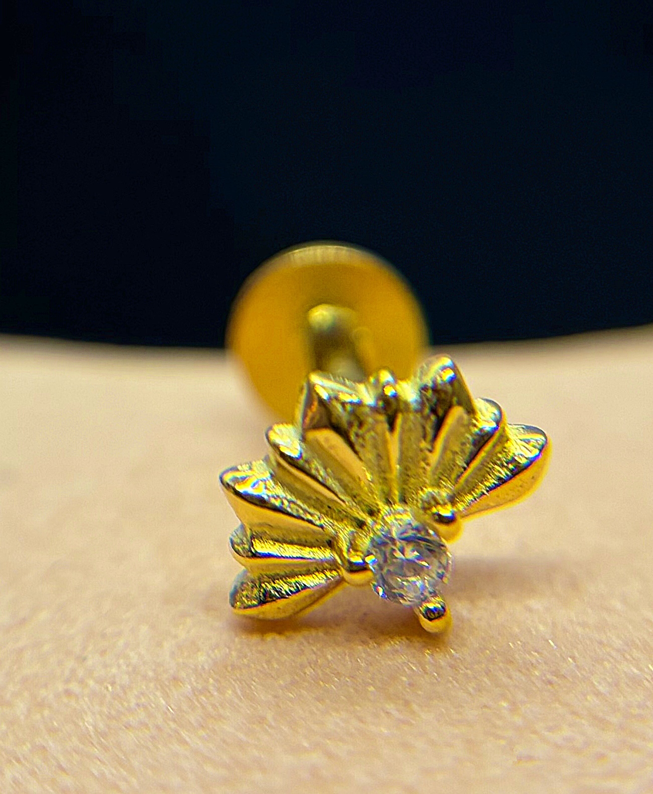Close-up of a small gold flower-shaped earring with a clear gemstone stud on skin against a dark background