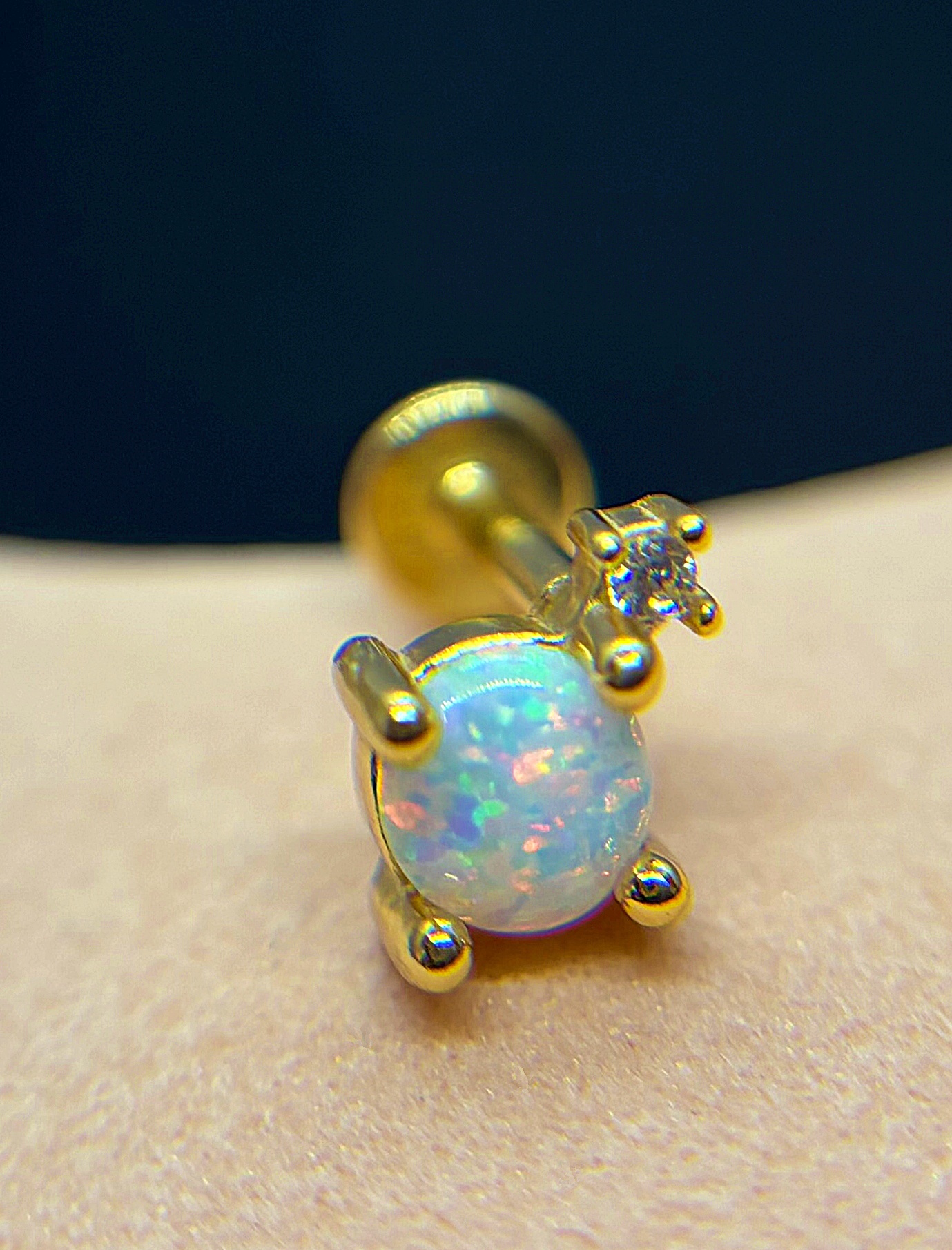 Close-up of a gold stud earring with a round opal showing blue-pink flashes and a small prong-set diamond accent