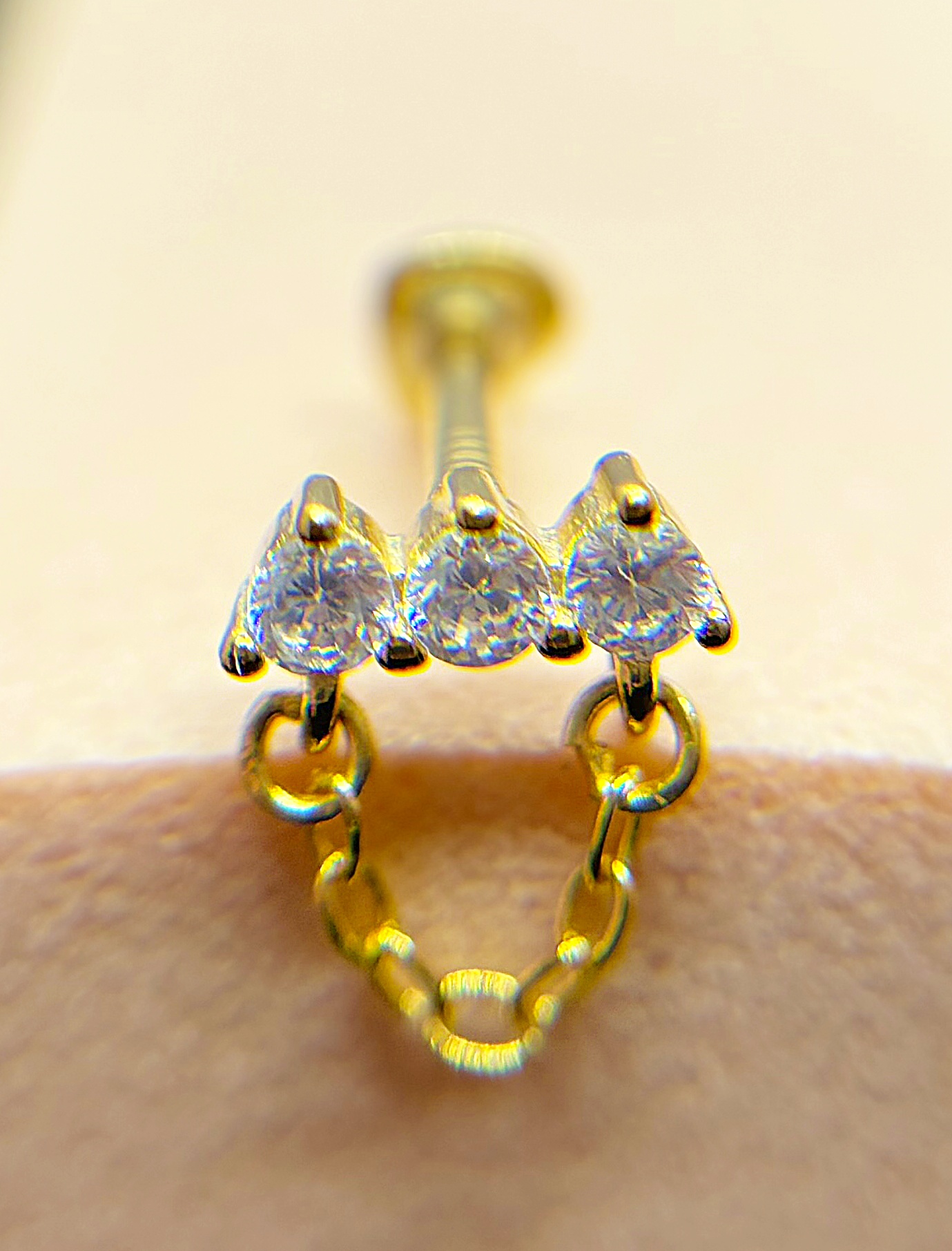 Macro close-up of a gold nose stud with three diamond-like stones and hanging chain detail on skin