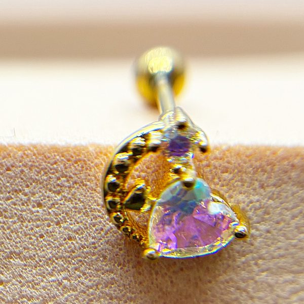 Close-up of a gold stud earring with a heart-shaped pink gemstone and twisted dotted gold detail on textured skin.