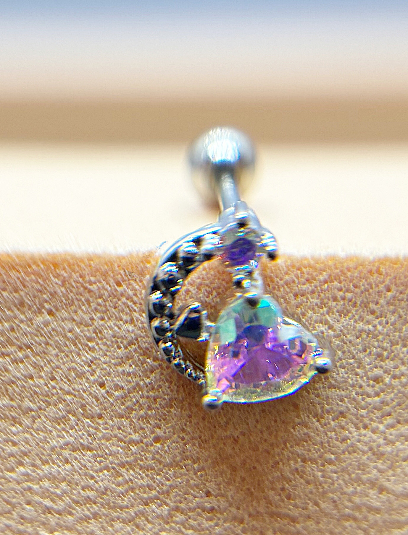Close-up of a silver curved stud earring with a heart-shaped iridescent purple gemstone set against textured skin.