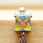 Close-up of a square iridescent gemstone earring on a tan surface, with a small dangling gem