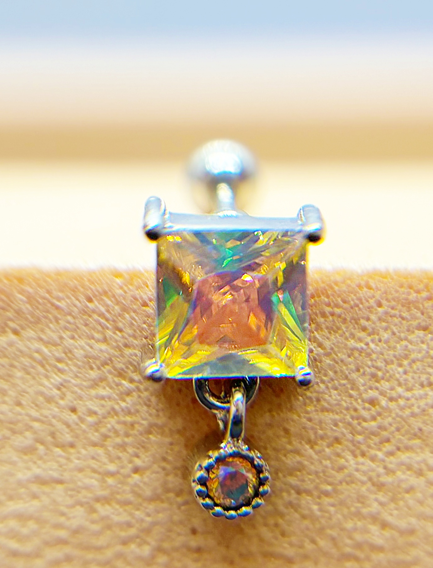 Close-up of a square iridescent gemstone earring on a tan surface, with a small dangling gem