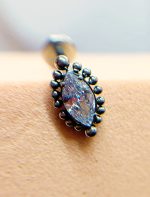 Close-up of a marquise-cut gemstone ring with a dark beaded setting on skin, with a blurred background necklace pendant behind it