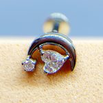 Close-up of a curved ear stud with two clear gemstones on skin, against a blue background