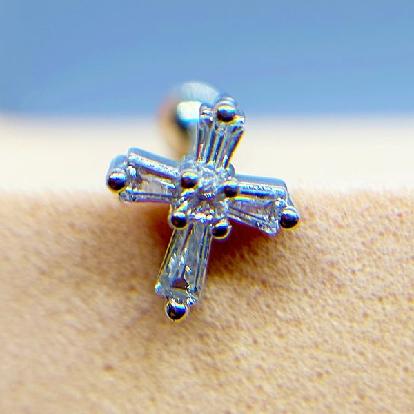 Close-up of a silver cross-shaped crystal earring against skin with a blue background