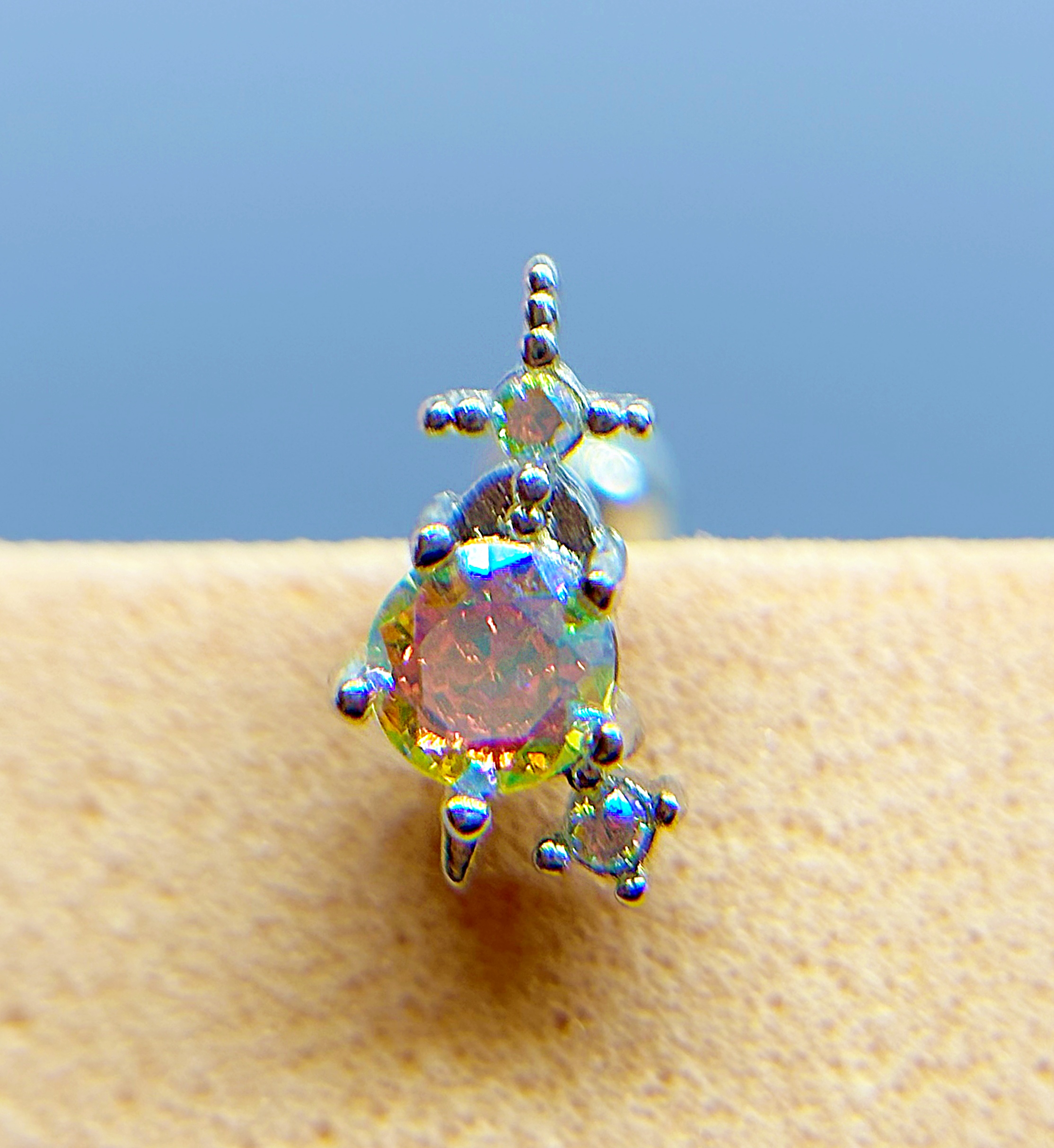 Close-up of an iridescent gemstone stud earring with a dangling accent on skin, against a blue background