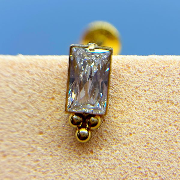 Close-up of a rectangular clear gemstone stud earring on a gold setting against a blue background