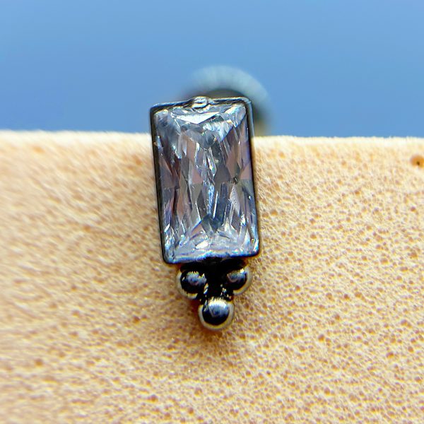 Close-up of a rectangular clear gemstone stud earring on skin, with a blue background