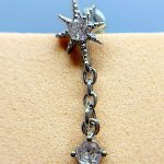 Close-up of a silver starburst earring with dangling chain and gem, worn on skin against a blue background