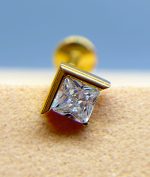 Close-up of a gold stud earring with a square clear gemstone against a blurred blue and beige background